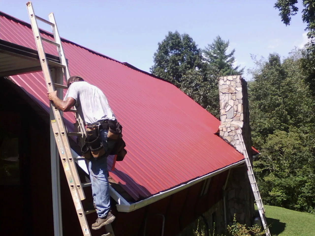 Roofer on a ladder beside a red metal roof and stone chimney — Soffit & Fascia Repair by Roofing Brothers in Genesee Depot
